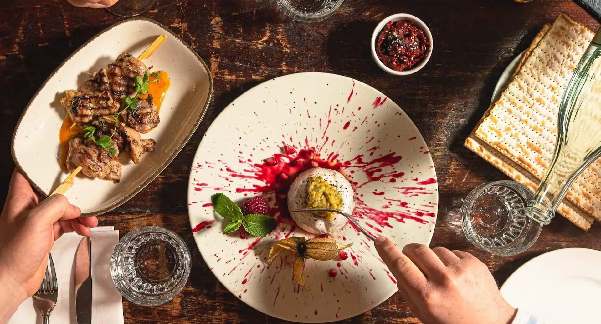Dessert auf mit Beerensauce bemaltem Teller, daneben Matze und Spieße